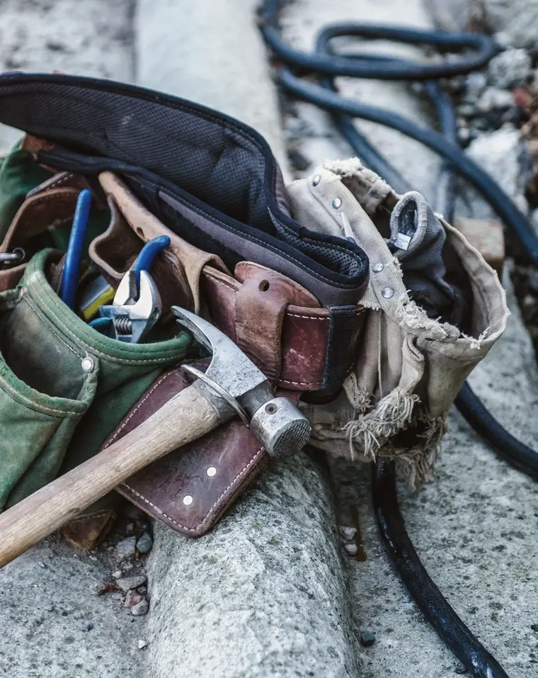 A worn leather and canvas tool belt with a hammer and pliers, resting on a stone ledge
